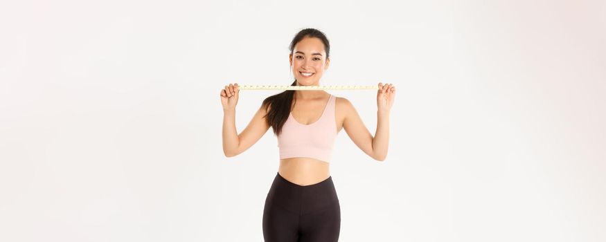 Fitness, healthy lifestyle and wellbeing concept. Portrait of smiling asian brunette sports girl, female athelte in activewear showing measuring tape, losing weight with exercises, white background