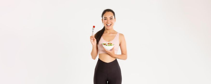 Sport, wellbeing and active lifestyle concept. Smiling slim and healthy asian girl doing fitness exercises, losing weight with gym workout and eating fresh salads, standing white background