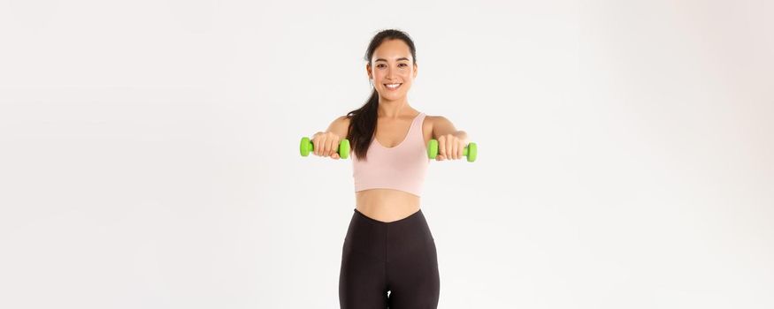 Sport, wellbeing and active lifestyle concept. Cheerful smiling asian fitness girl, sportswoman lifting dummbells, workout on muscles, gaining biceps with home exercises, white background