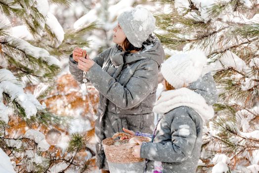 Mother and daughter in winter time in snow forest decorate Christmas tree outdoors. Girl with child kid in wood in cold weather