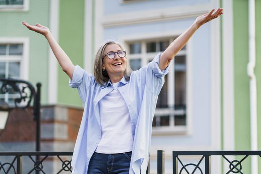 Mature grey hair woman wearing glasses standing outdoor traveling around world at retirement. Senior woman meeting her family. Mature woman with perfect skin wearing blue shirt standing outside