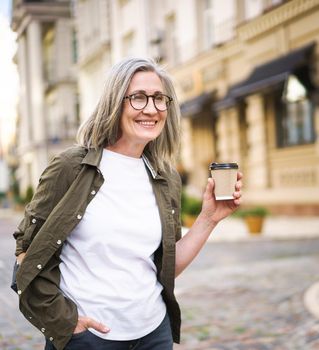 Happy mature businesswoman with disposable cup of hot coffee in hand. Charming senior woman traveling in Europe enjoying cup of tea, coffee. Grey haired woman in casual with backpack