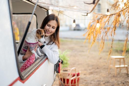 Caucasian woman looks out of the window of the van while hugging her dog Jack Russell Terrier. Travel in a camper in autumn