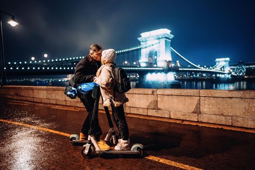 Young happy couple kissing with electric scooter outside in front of ancient european bridge in winter.
