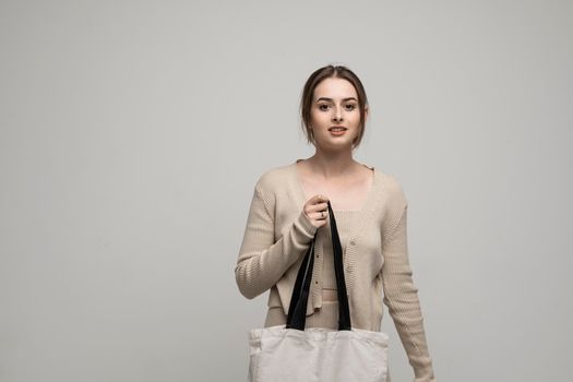 Beautiful brunette woman in a beige costume with cotton bag on in a hand on a grey background. Mockup and zero waste concept.