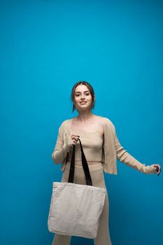 Beautiful brunette woman in a beige costume with cotton bag on in a hand on a blue background. Mockup and zero waste concept.