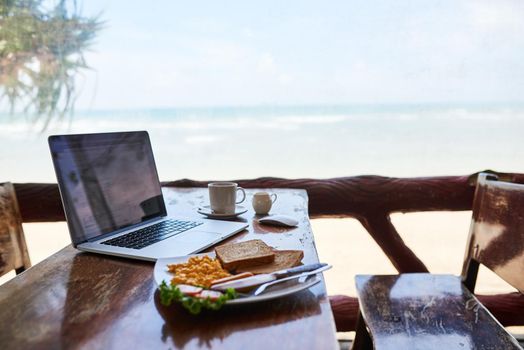 Breakfast with the best view. a laptop and freshly made breakfast on a table with a view of the beach in the background.