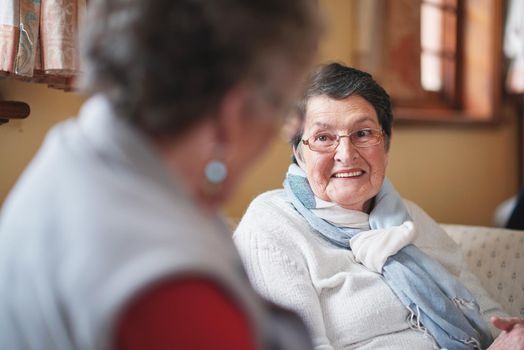 Happy elderly woman talking to friend sitting on sofa in retirement home having conversation