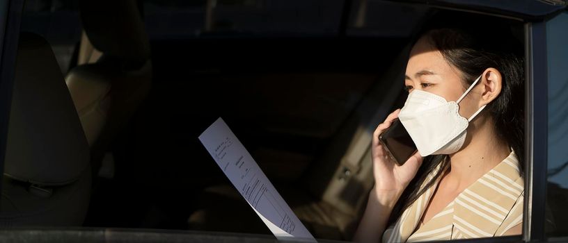 Smiling businesswoman wearing protective face mask and talking on mobile phone while sitting on back seat in the car.