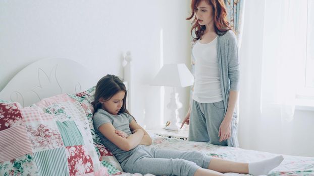 Angry mother argue scolding her upset little daughter lying on bed in bright bedroom at home