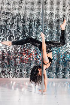 Woman doing tricks, practicing in studio with pylon. Young stripper in black dances sexually with pole in club. Poledance on shining wall background