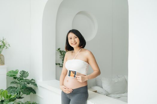 Cheerful pregnant woman holds ultrasound photo at her belly