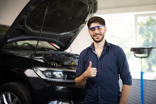 Happy technician bearded brunet mechanic man 20s in safety glasses work in vehicle repair shop show thumb up like gesture on light workshop background indoors