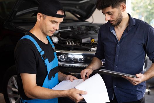 Car Service Employees Inspect the Bottom and Skid Plates of the Car. Manager Checks Data on a Tablet Computer and Explains the Breakdown to Mechanic. Specialist is Showing Info on Tablet.