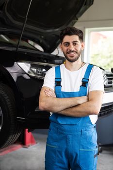 Male car mechanic at workplace in spacious repair shop. Portrait of bearded car mechanic crosses hands in a car workshop in blue uniform with equipment looking into camera