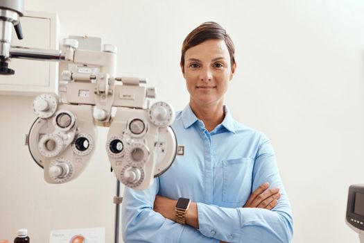 Confident, happy and proud optometrist standing with arms crossed, ready for checkup and preparing equipment in an optometry office. Smiling, successful and wellness portrait of an ophthalmologist