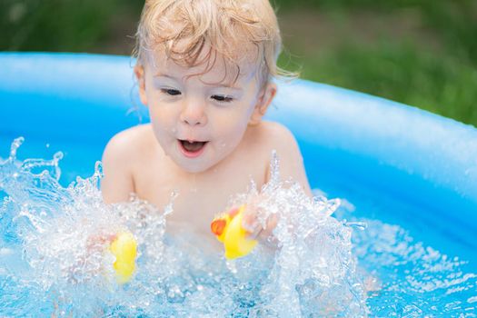 Cute little child bathing with duck in blue street pool in courtyard. Portrait of joyful toddler, baby. Kid laughs, splashes water, smiles. Concept of healthy lifestyle, family, leisure in summer