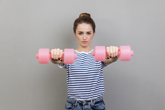 Serious concentrated woman holding out pink dumbbells to camera, work out alone.