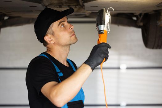Auto mechanic man in blue overalls with lamp working at workshop. Professional mechanic is working on a car in a car service. Car service, repair, maintenance and people concept