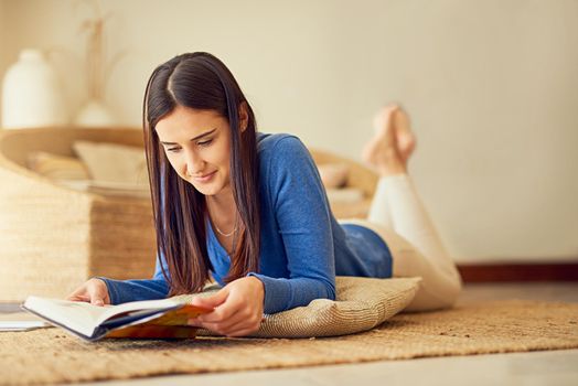 Making time for herself this weekend. a happy young woman reading a book on her lounge floor at home.