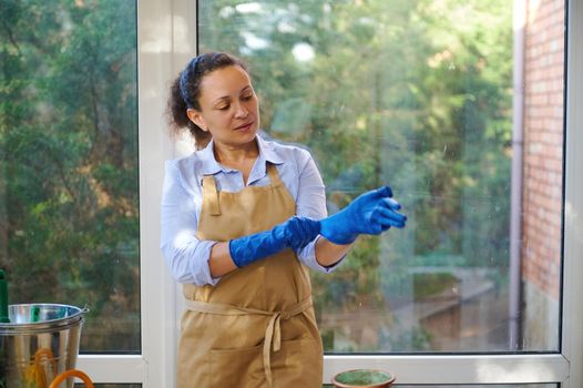 Multi-ethnic woman florist, gardener, horticulturist in beige apron putting on rubber work gloves, at home terrace