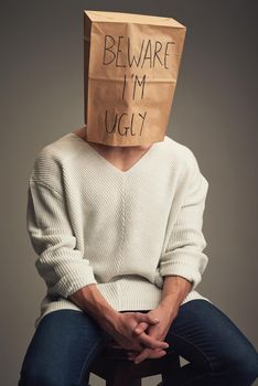 They think Im ugly but Im not. an unrecognizable man wearing a paper bag saying beware Im ugly while being seated against a grey background.