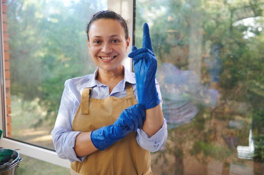 Pretty housewife, maid, florist, horticulturist in beige apron puts on blue rubber work gloves, smiles looking at camera
