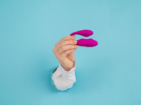 A woman's hand holds a pink sex toy through a hole in a paper blue background.