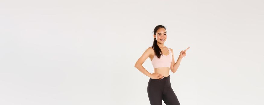 Sport, gym and healthy body concept. Full length of smiling beautiful asian sportswoman in sportswear, pointing finger left and showing advertisement, gym membership or sports equipment