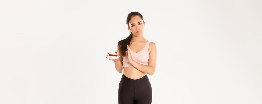 Active lifestyle, fitness and wellbeing concept. Determined asian girl athlete rejecting sweets, quit eating junk food during diet, losing weight, refuse eat cake, standing reluctant white background