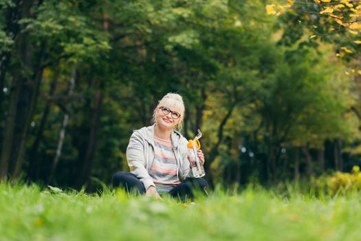 Senior beautiful woman sitting in the park on the grass and drinking fresh juice, water after sports, yoga