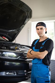 Portrait shot of caucasian man in blue uniform and hat standing in big auto garage, looking to camera and crossing hands. Pretty male mechanic in maintaining service