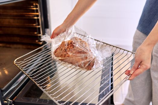 Hands woman cooking pork at home in baking sleeve, puts tray of meat in oven