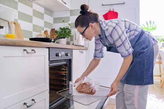 Woman preparing pork in baking sleeve at home, puts tray of meat in oven
