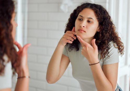 Woman popping a zit during a skincare routine while in front of a mirror in the bathroom. Young Unhappy, upset or annoyed lady with a pimple targeting acne, dry skin or pimple on her face at home