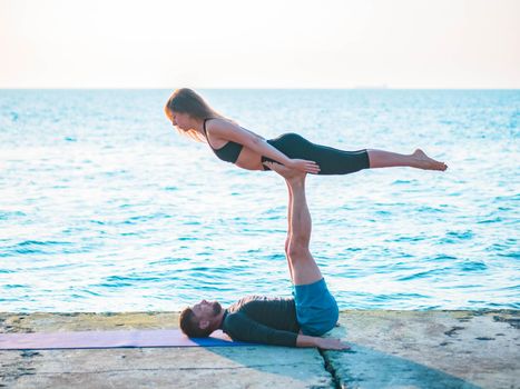 Fit young couple doing acro-yoga at sea beach. Man lying on concrete plates and balancing woman on his feet. Beautiful pair practicing yoga together.