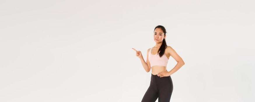 Sport, gym and endurance concept. Full length of skeptical and unamused female athlete, sportswoman in active wear looking reluctant, smirk and pointing finger left with judgement, white background