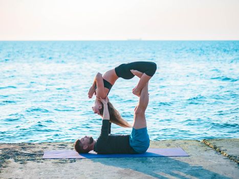 Young beautiful couple practicing acro yoga on the sea beach near water. Man and woman doing everyday practice outdoor on nature background. Healthy lifestyle concept.