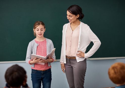 Shes a confident speaker. an elementary school girl reading in front of the class while standing alongside her teacher.