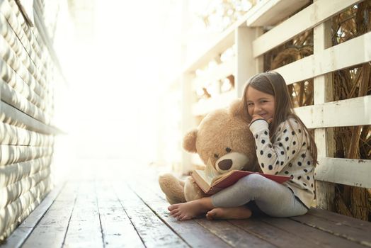 The sunshine days of childhood. Portrait of a little girl reading a book with her teddy bear beside her.