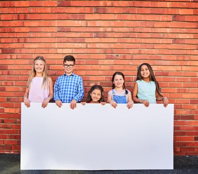 Well use our cuteness to help promote your message. Portrait of a group of young children holding a blank sign against a brick wall.