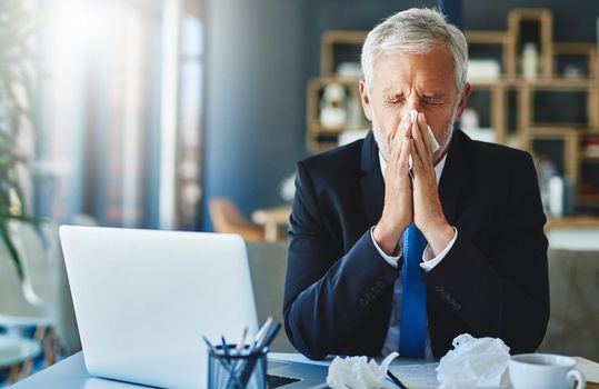 I cant afford to be sick now. a frustrated businessman using a tissue to sneeze in while being seated in the office.