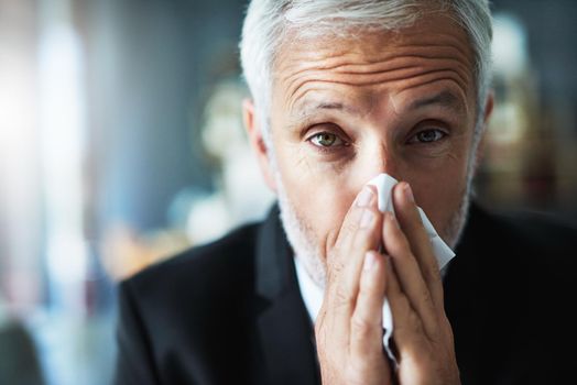 I have to remain positive. a frustrated businessman using a tissue to sneeze in while being seated in the office.