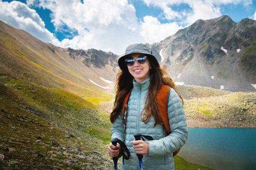 Smiling, positive girl with disheveled hair against the background of a blue mountain lake, with trekking poles in her hands and a backpack, a tourist on a hike