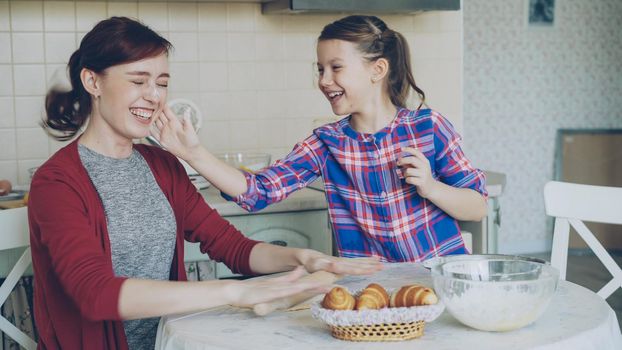 Happy mother and cute daughter have fun smearing nose each other with flour while cooking together in the kitchen on holidays. Family, food, home and people concept