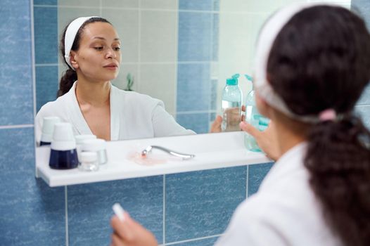 Charming middle-aged woman in white bathrobe, standing in front of a bathroom mirror and holding a face cleanser