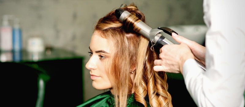 Hairdresser using curling tongs curls long brown hair on the young caucasian girl in a beauty salon.