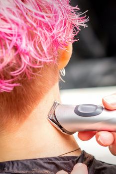 Back view of hairdresser's hand shaving nape and neck with electric trimmer of young caucasian woman with short pink hair in beauty salon.