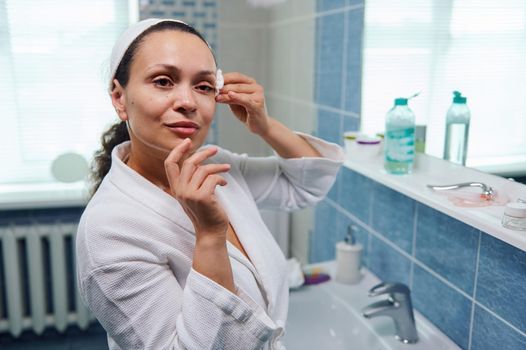 Beautiful multi-ethnic woman in bathrobe, using a cotton pad, cleans her face with a face cleanser and removes make-up