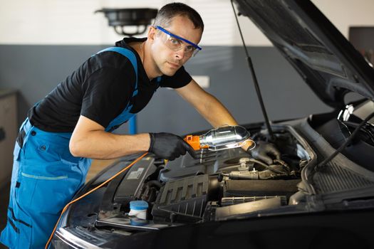 Portrait of young caucasian man in blue overalls and safety glasses inspects engine with flashlight. Male car mechanic at work in spacious repair shop. Modern workshop
